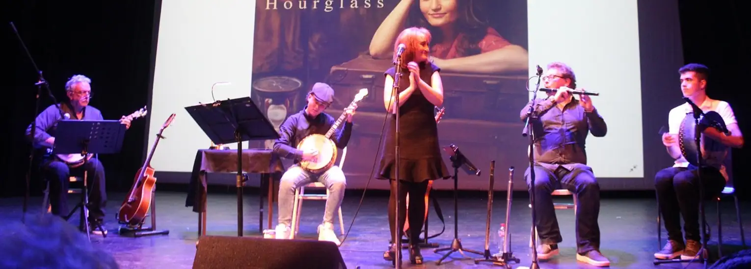 A band performs on stage with a singer at the centre, flanked by musicians playing banjo, guitar, flute, and accordion. A large projected image of a woman and the word “Hourglass” appears behind them.