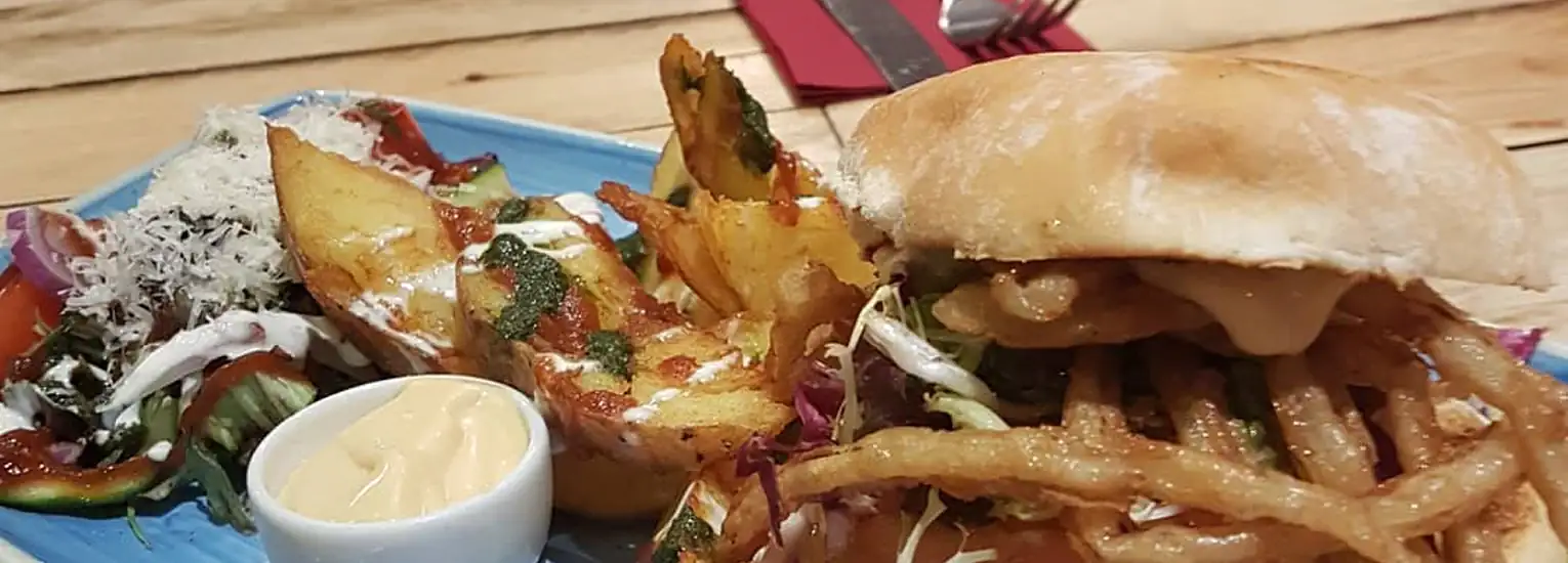 A blue plate with a sandwich filled with crispy fried onions, served with thick potato wedges, a side of mayonnaise dip, and a fresh salad. Eating utensils and a red serviette are visible in the background.