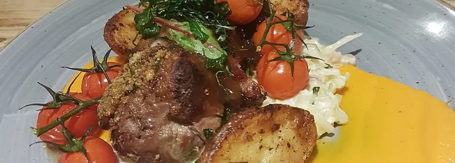 A plate of roasted meat topped with herbs, surrounded by crispy roast potatoes, cherry tomatoes on the vine, a bed of coleslaw, and a smear of orange sauce on a grey plate.