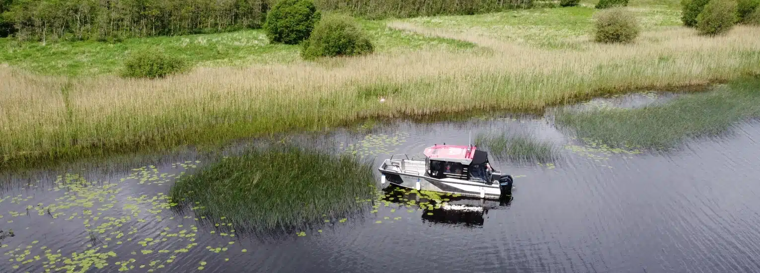 A small motorboat with a red roof floats on a calm river surrounded by tall grass, reeds, and green vegetation, with lily pads scattered on the water's surface.