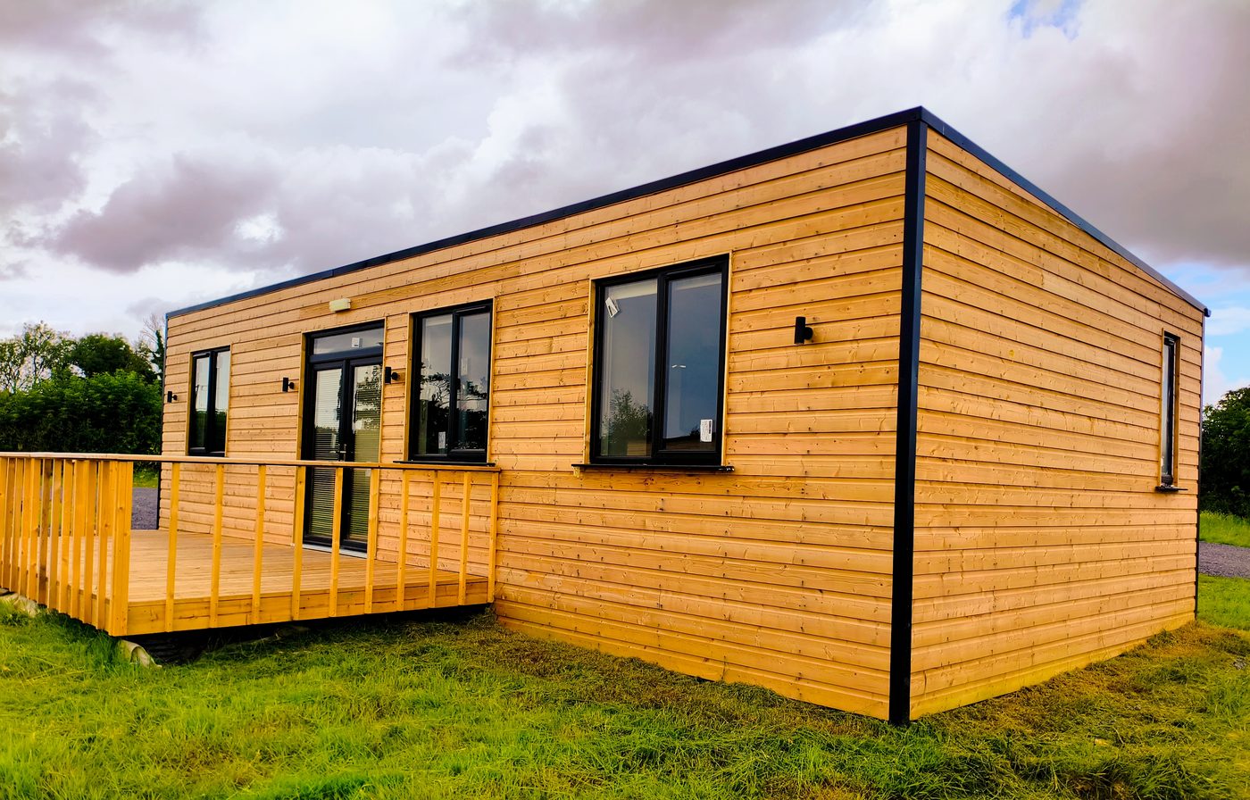 A modern tiny house with wooden exterior cladding, large windows, and a small front veranda sits on green grass under a cloudy sky.